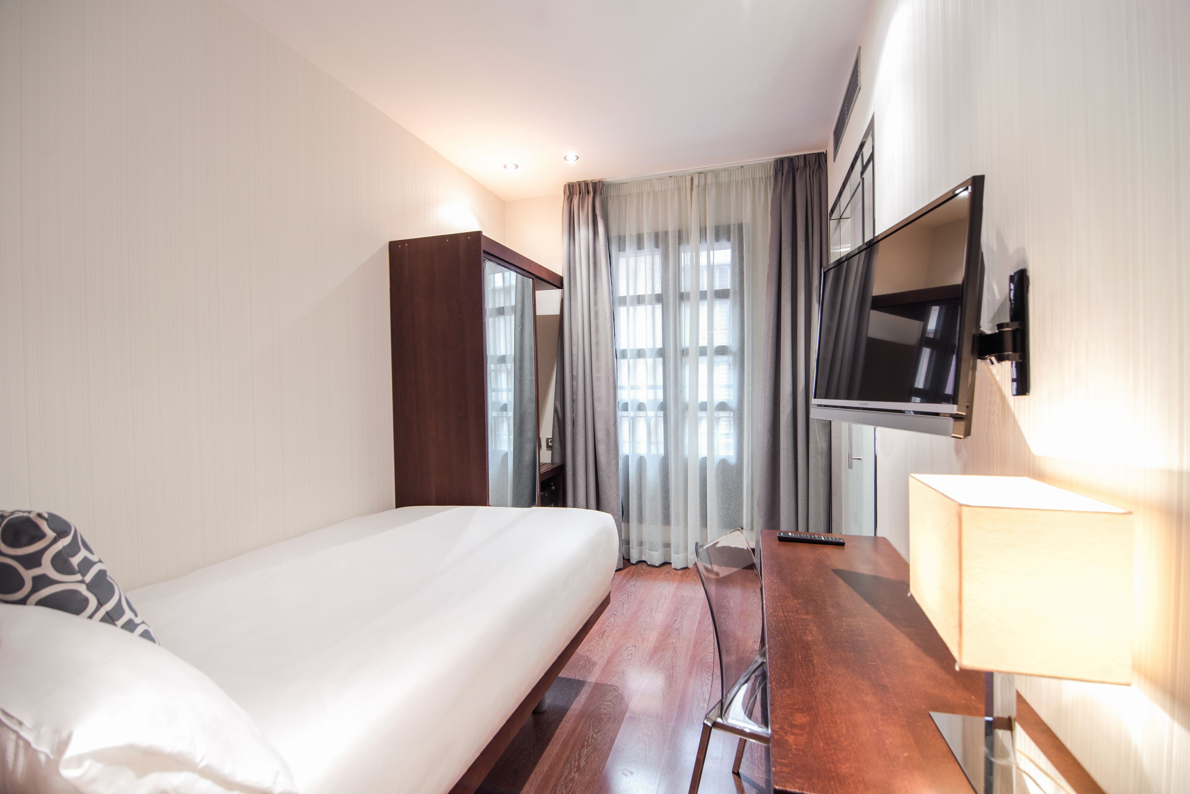 Habitación de hotel con cama, escritorio, televisor montado en la pared y ventana con cortinas.