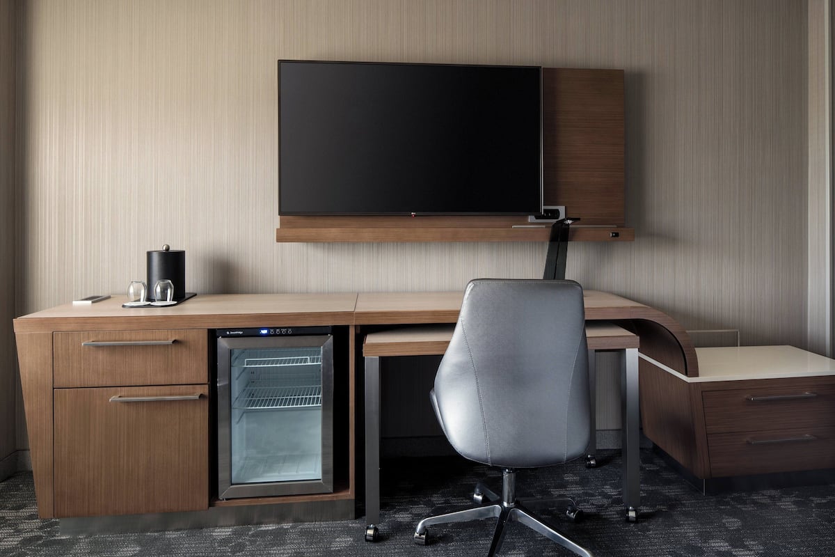Une chambre d’hôtel avec un bureau en bois, un téléviseur à écran plat fixé au-dessus, un mini-réfrigérateur, un fauteuil de bureau gris et un four à micro-ondes encastré.