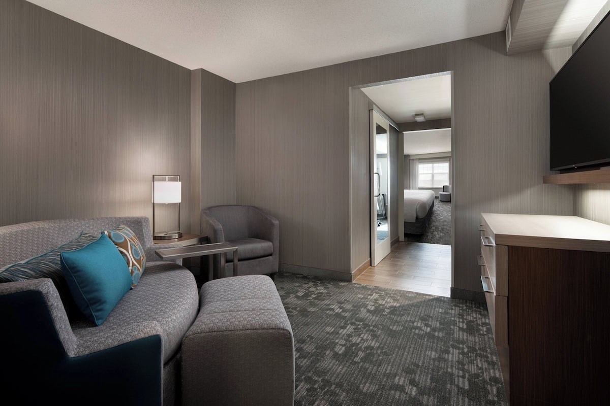 Une chambre d’hôtel moderne avec un canapé gris, une petite table avec une lampe, un téléviseur fixé au mur et un meuble en bois.