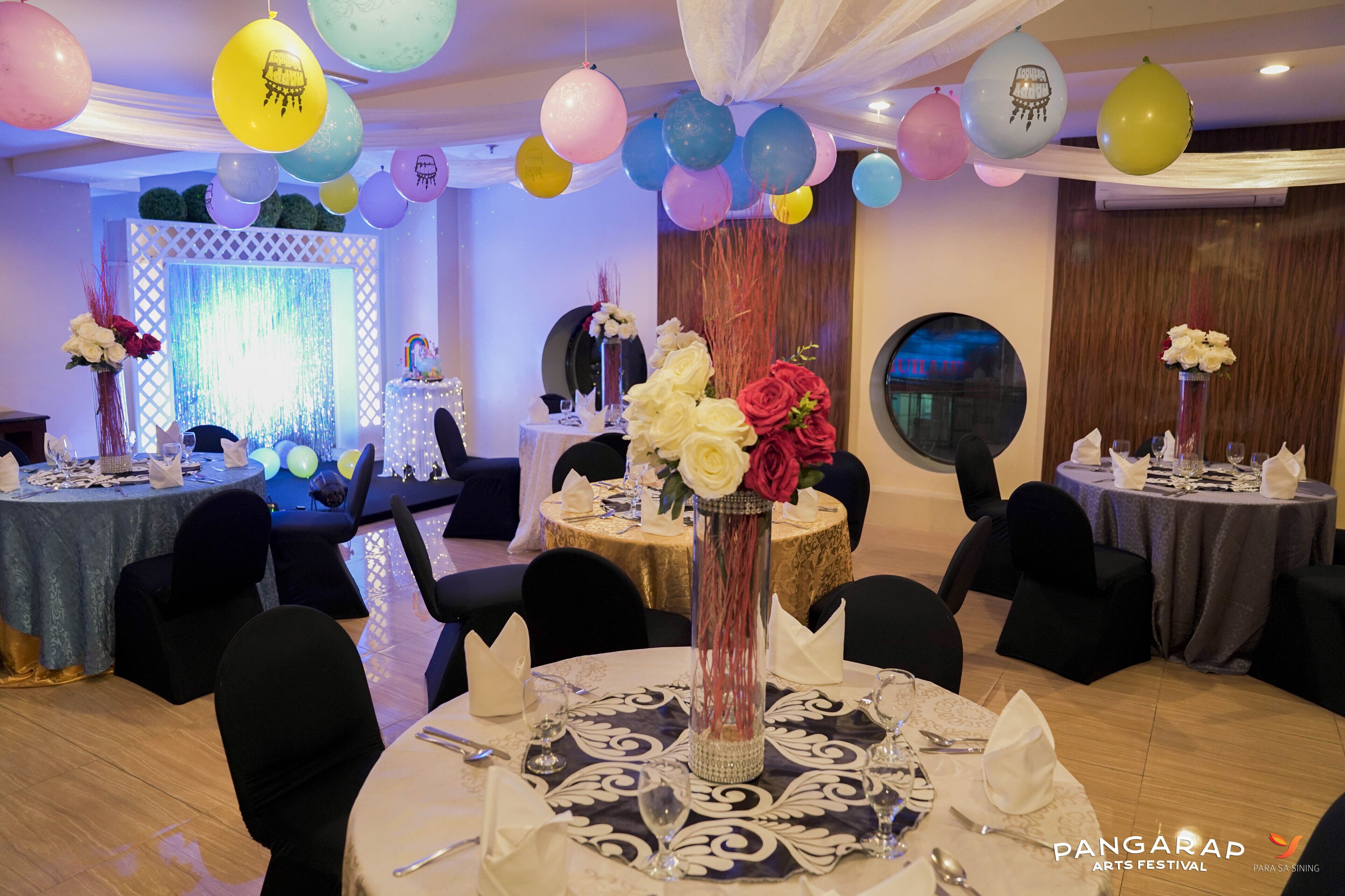 A banquet hall with round tables, black chairs, and a stage with a backdrop.