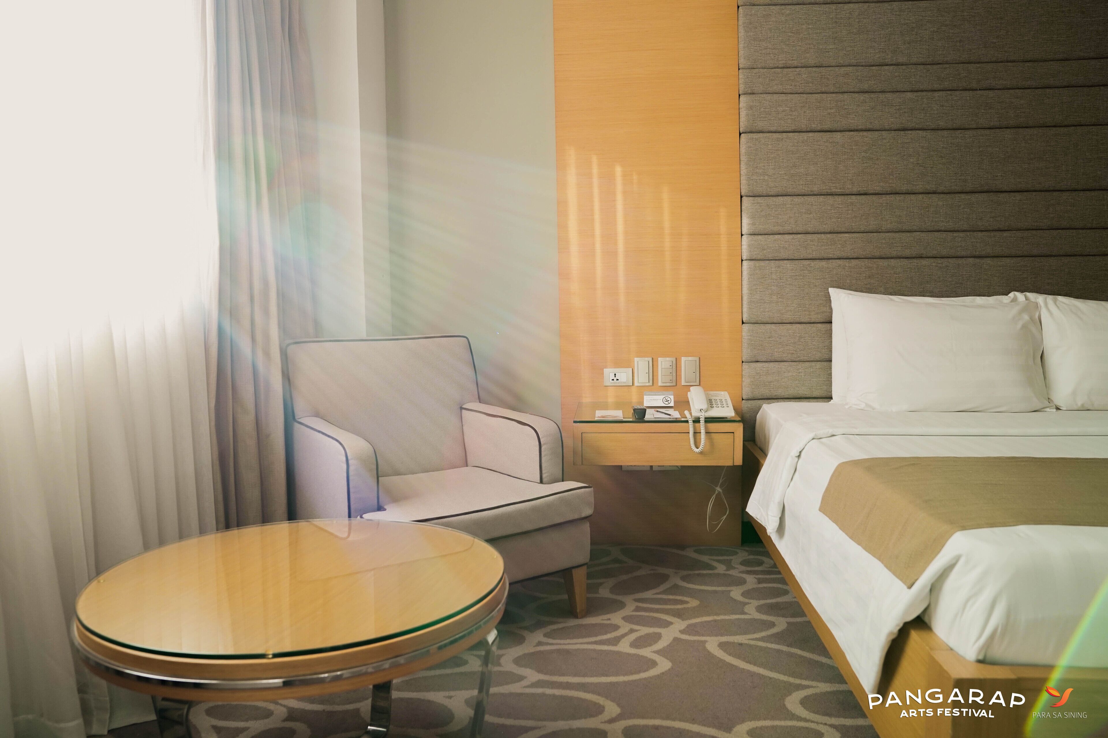 A hotel room with a bed, a round glass table, a chair, a nightstand with a phone, and a wall-mounted light fixture.