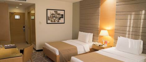 A hotel room with two beds, a desk, and a chair. There is a framed picture on the wall and a lamp on the nightstand.