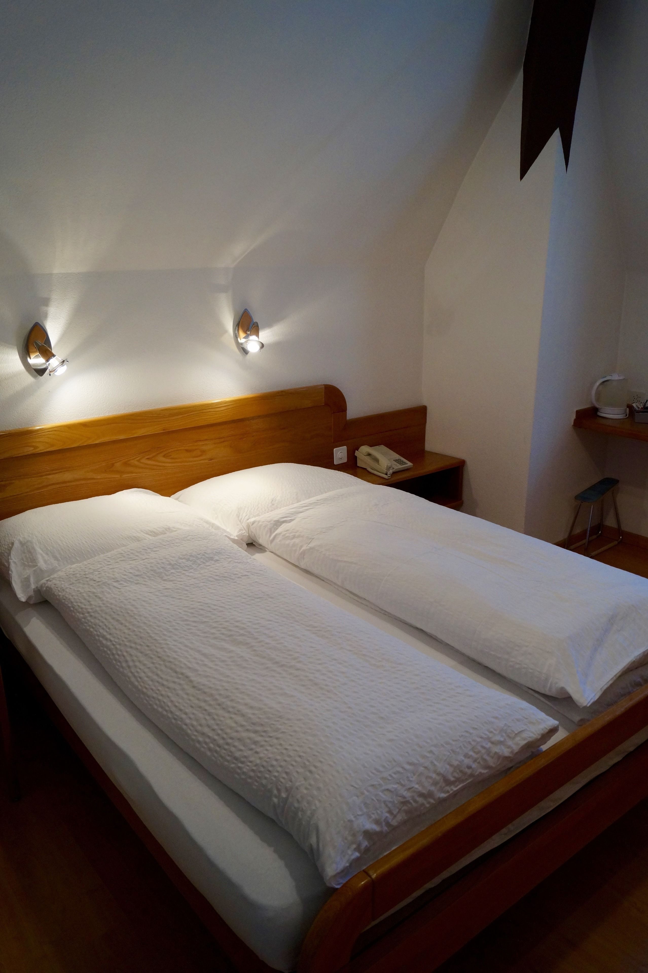A neatly made bed with a wooden headboard, two bedside lamps, a bedside table with a phone, and a small shelf with a cup.