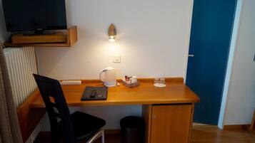 Habitación compacta con escritorio de madera, una silla, televisor montado en la pared, estantería pequeña, luz en la pared y una puerta.