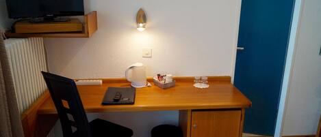 Habitación compacta con escritorio de madera, una silla, televisor montado en la pared, estantería pequeña, luz en la pared y una puerta.