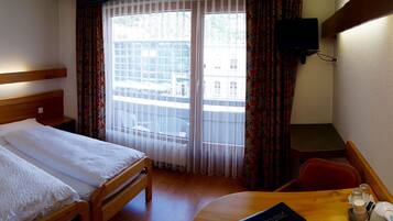 Quarto de hotel com cama, escrivaninha, cadeira, televisão e janela com cortinas.