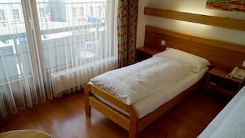 A single bed with a wooden frame, a bedside table with a phone, and a window with sheer curtains.
