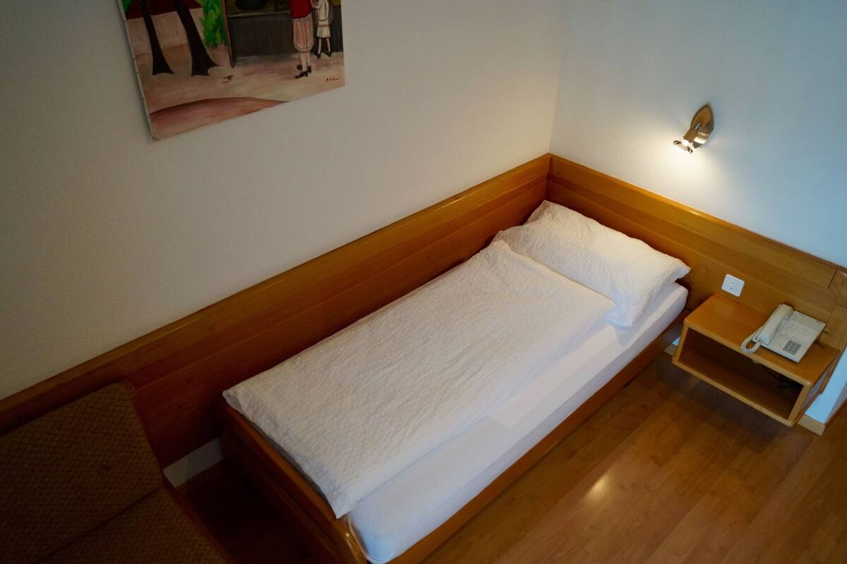A single bed with a wooden headboard, a bedside table with a telephone, and a wall-mounted light fixture.