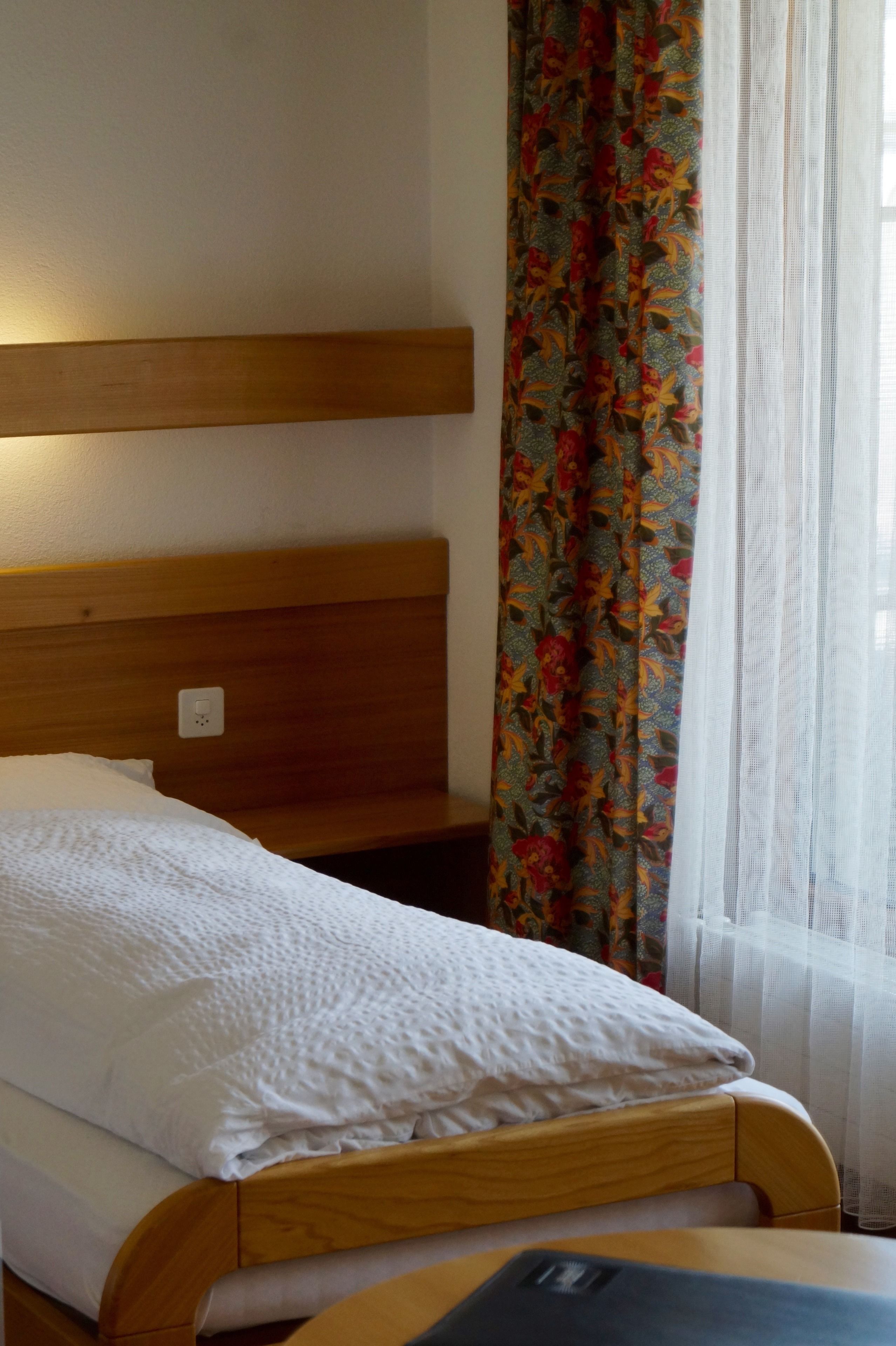 Una cama individual con cabecera de madera, almohada y manta blancos.  Junto a la cama, una mesita de noche de madera. Una cortina floral de colores cubre parcialmente una ventana.