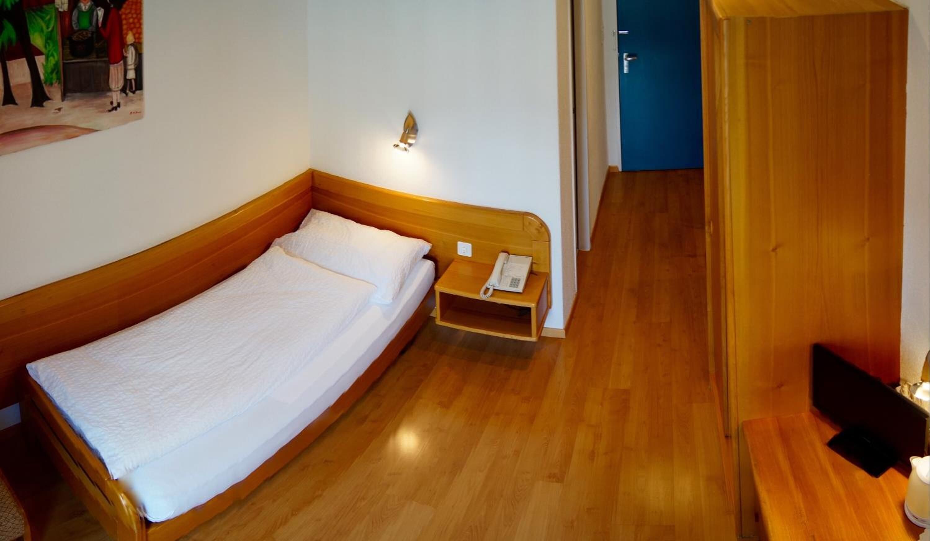 Un lit simple avec une tête de lit en bois, une table de chevet avec un téléphone et un luminaire fixé au mur.