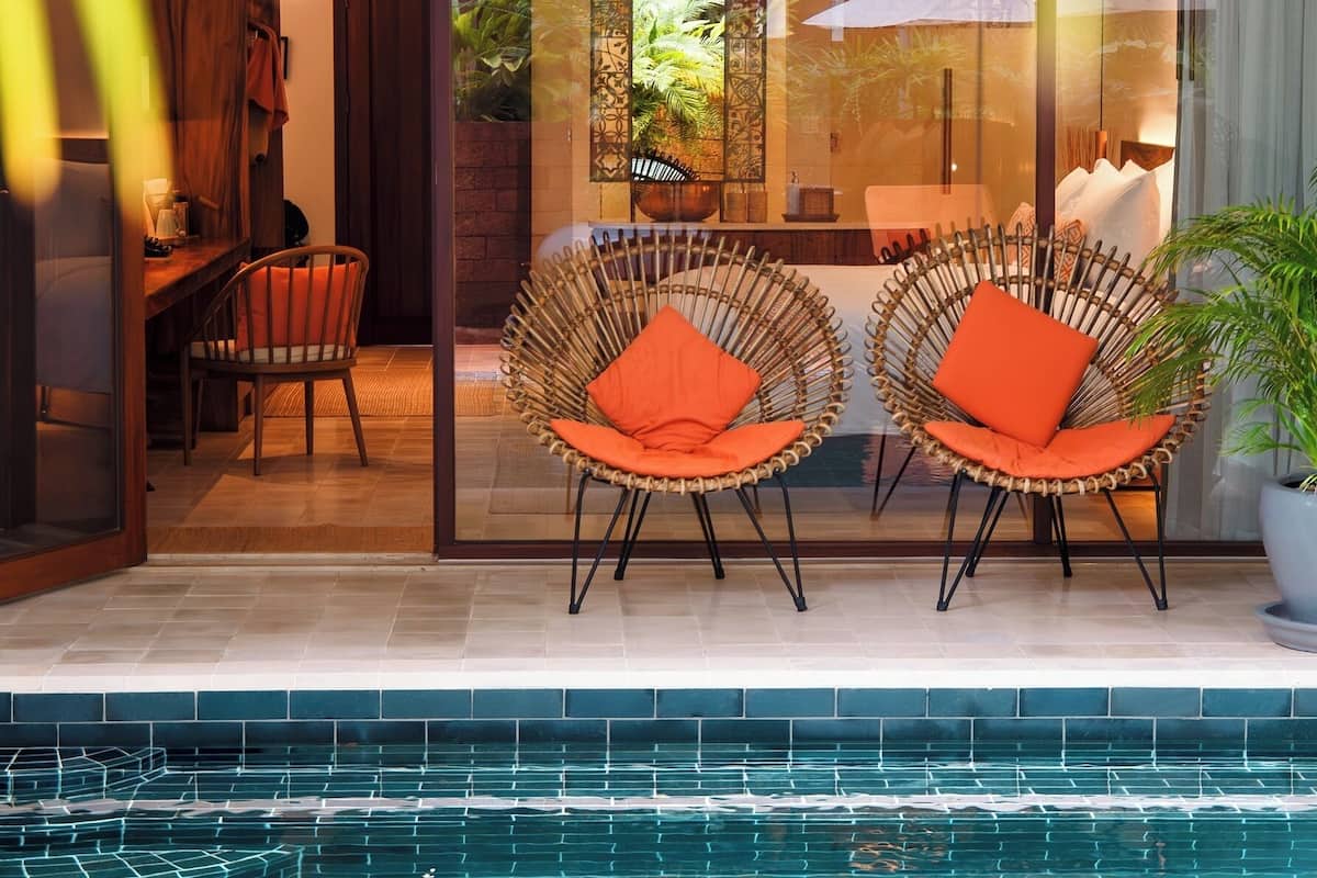 A poolside area with wicker chairs and a table, a glass door leading to an indoor space, and a ceiling fan.