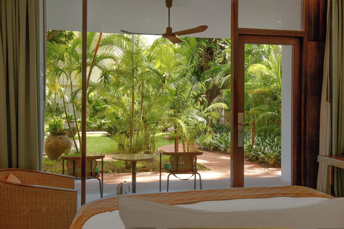 A bedroom with a large window, a ceiling fan, and a view of a lush garden.
