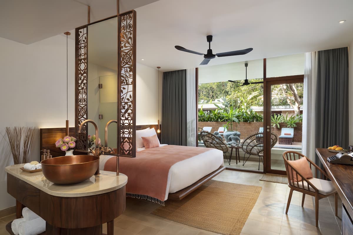A modern hotel room with a large bed, a sitting area with chairs, a desk, and a view of the outdoors.