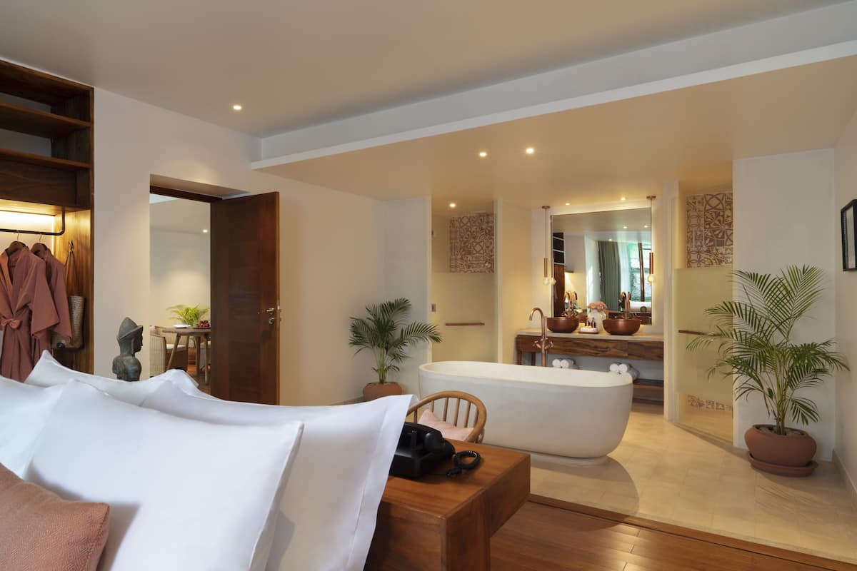 A modern hotel room with a large bed, a wooden desk, a bathtub, and a bathroom with a sink and mirror.