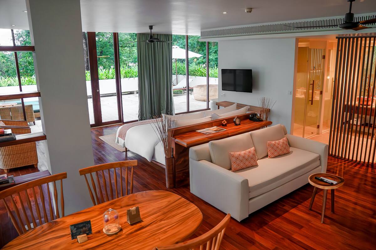A modern hotel room with a bed, sofa, dining table, and a TV.