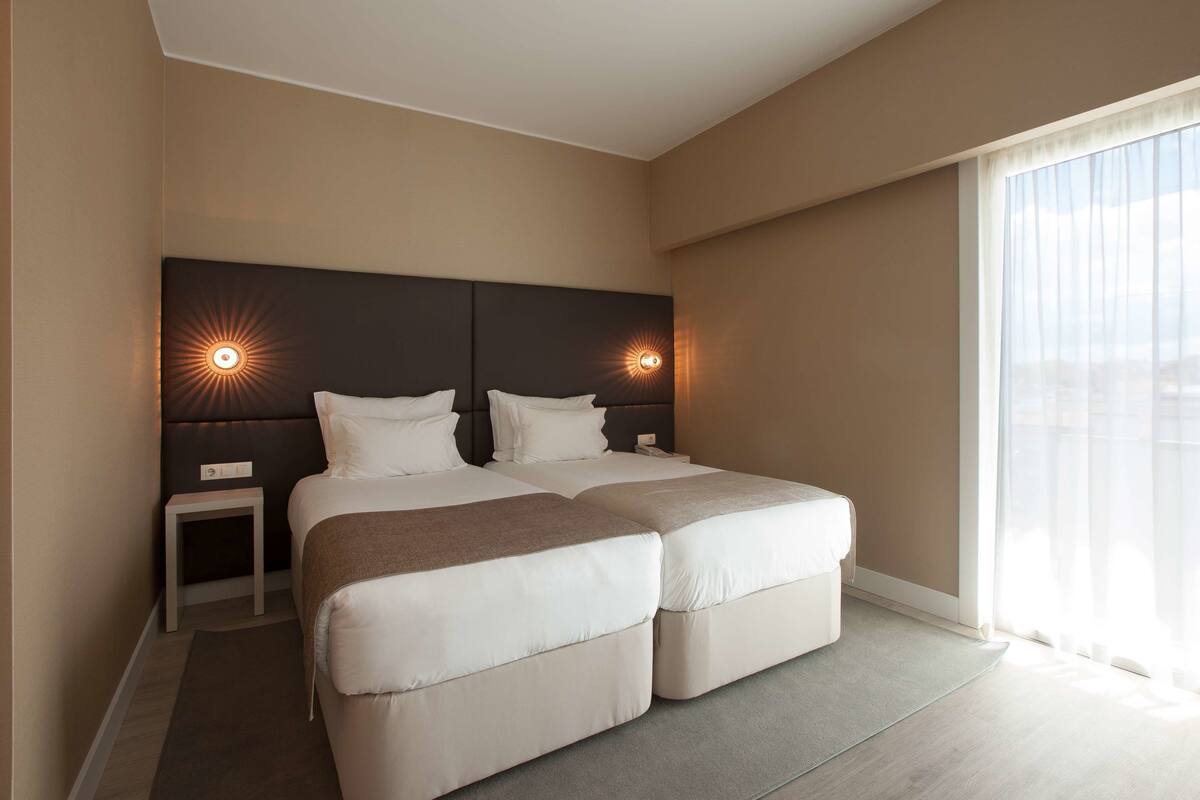 A modern hotel room with two beds, a small bedside table, and a large window with sheer curtains.