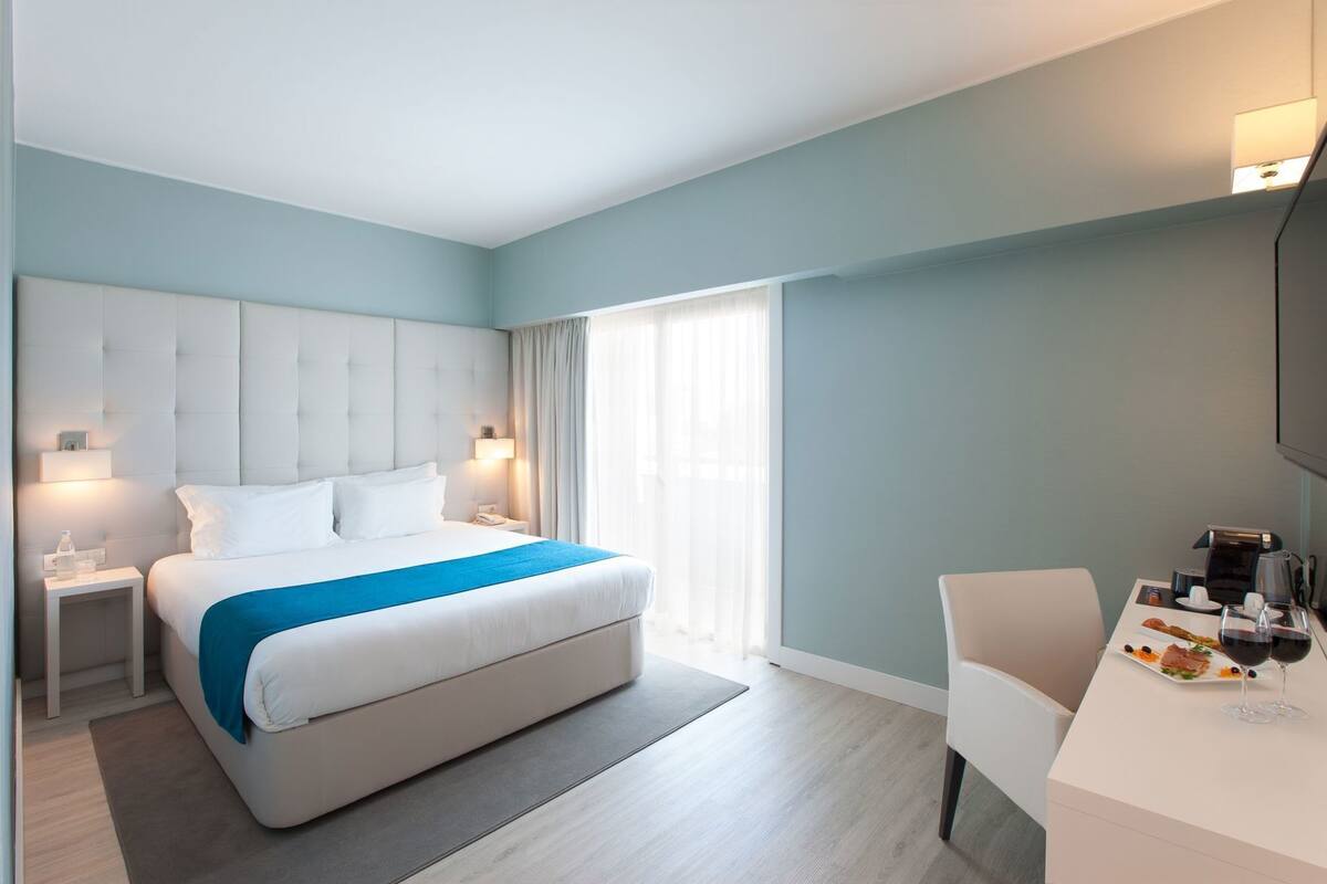 A modern hotel room with a large bed, bedside tables, a desk with a chair, and a television.