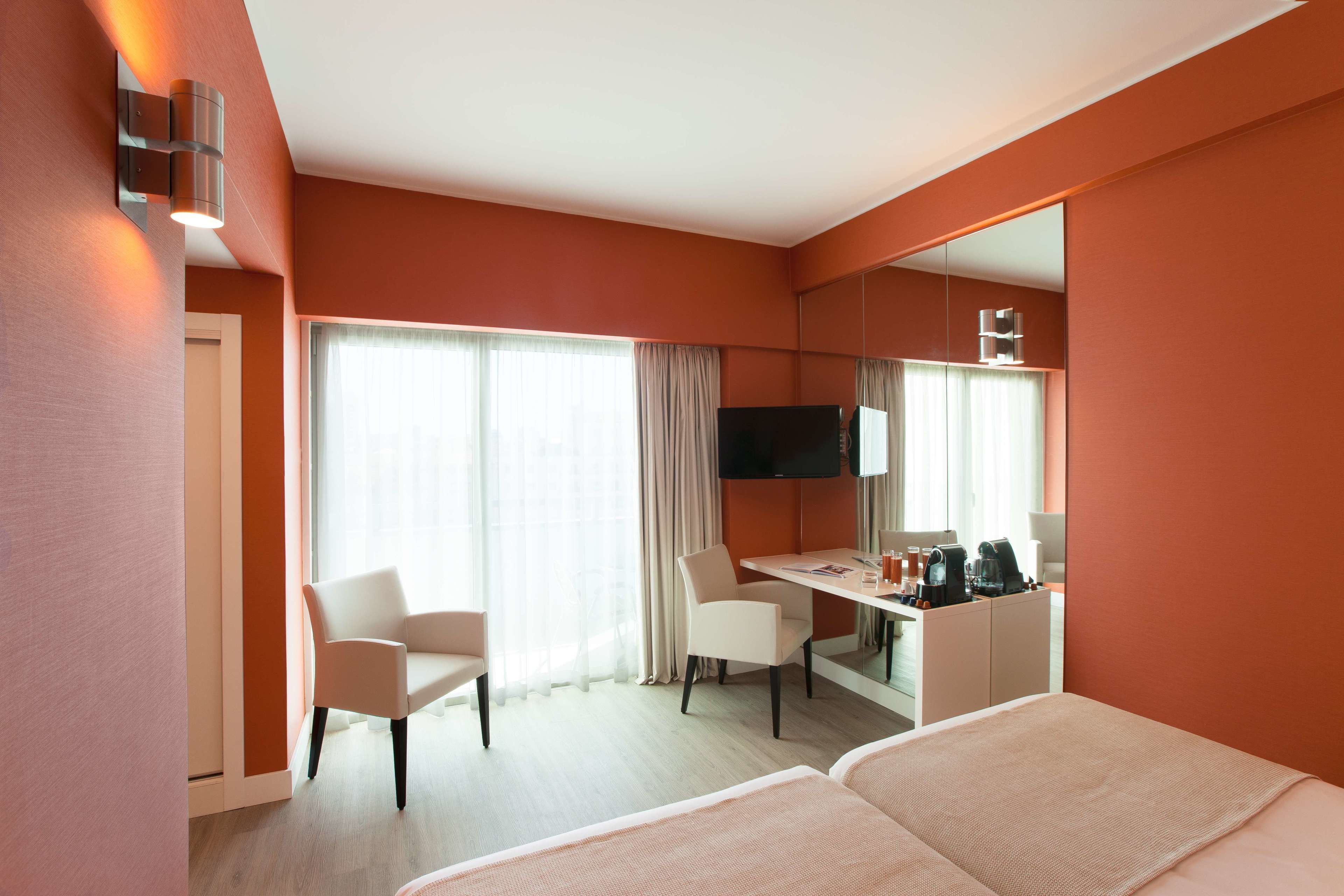 Habitación de hotel con cama, escritorio, silla, televisión y un amplio ventanal con cortinas.