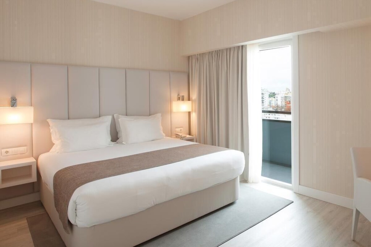 A modern hotel room with a large bed, bedside lamps, a view to the outside, and a balcony.