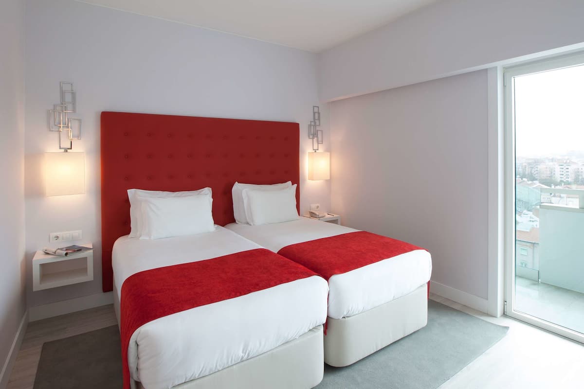 A hotel room with a red headboard, two beds, bedside tables with lamps, and a large window offering a city view.