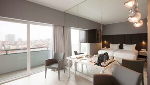 Una habitación de hotel moderna con un balcón grande, un televisor de pantalla plana, una cama con cabecera oscura y una zona de comedor con cafetera.