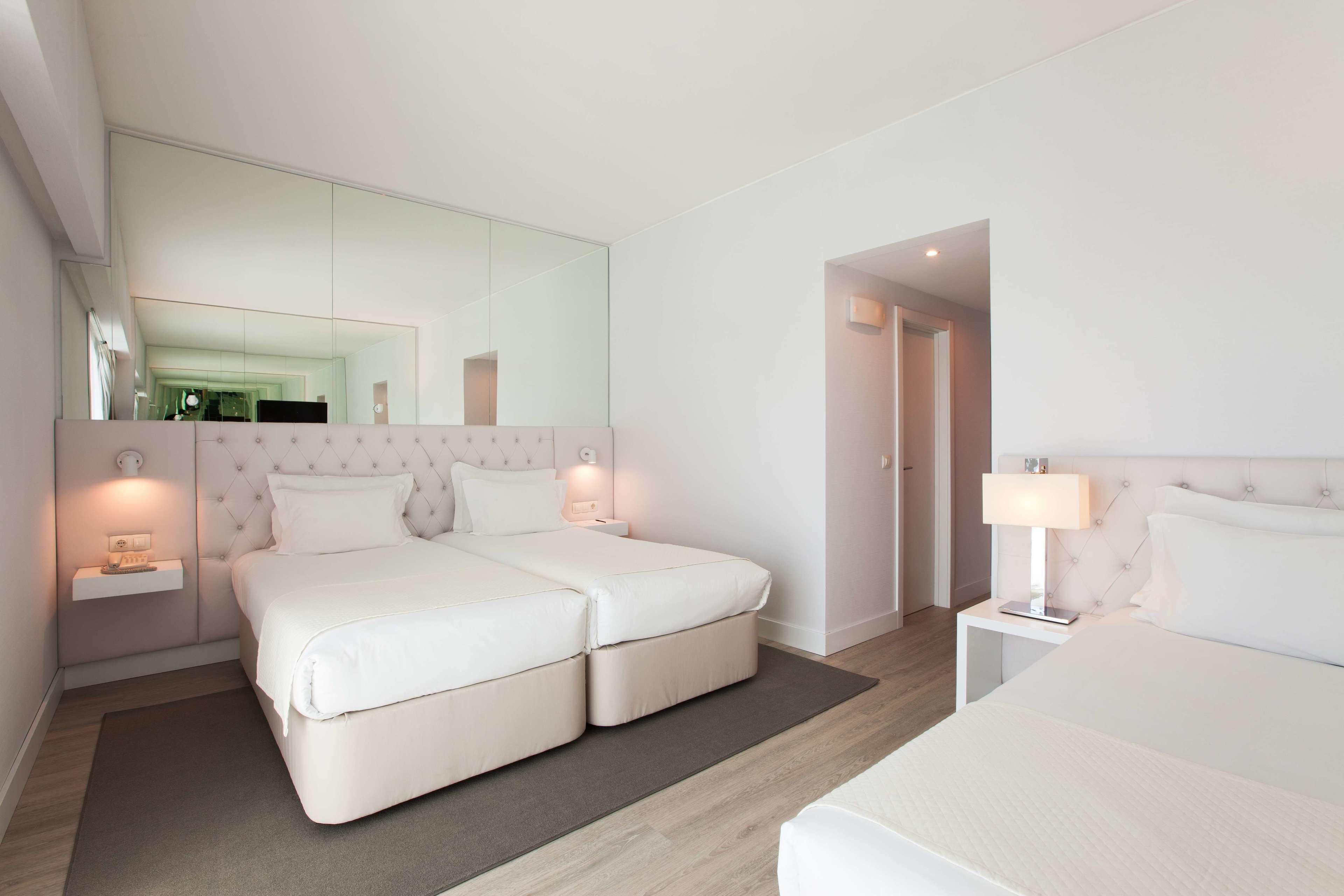 Una habitación de hotel moderna con una cama grande, mesitas de noche, un espejo y una puerta que da a otra habitación.