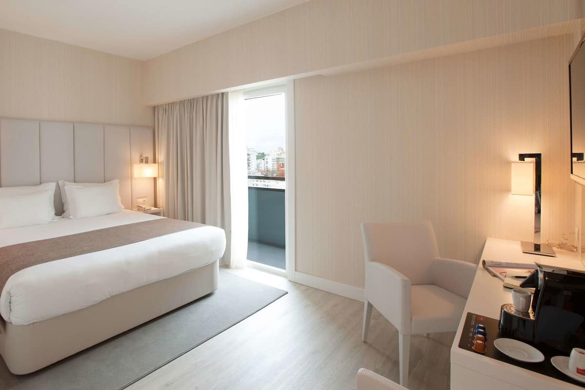 A hotel room with a large bed, a desk with a chair, a coffee machine, and a view of the city through the window.