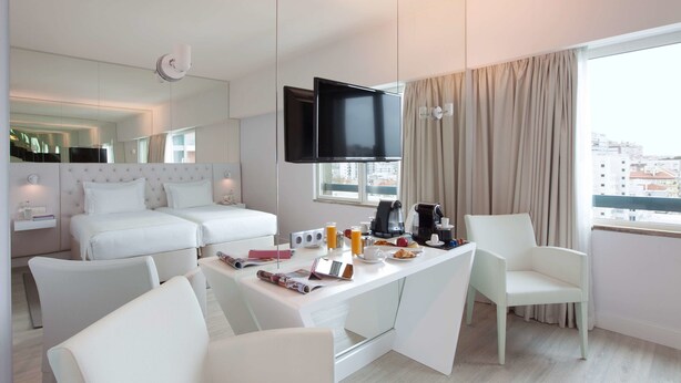 Habitación de hotel moderna con zona de comedor, cama con ropa blanca, televisor de pantalla plana y vista a la ciudad.