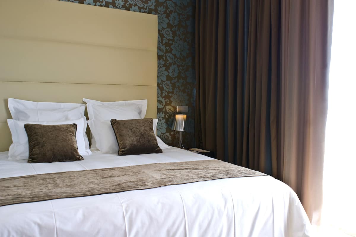 A neatly made bed with white linens and two brown pillows, a headboard, and a wall with floral wallpaper.
