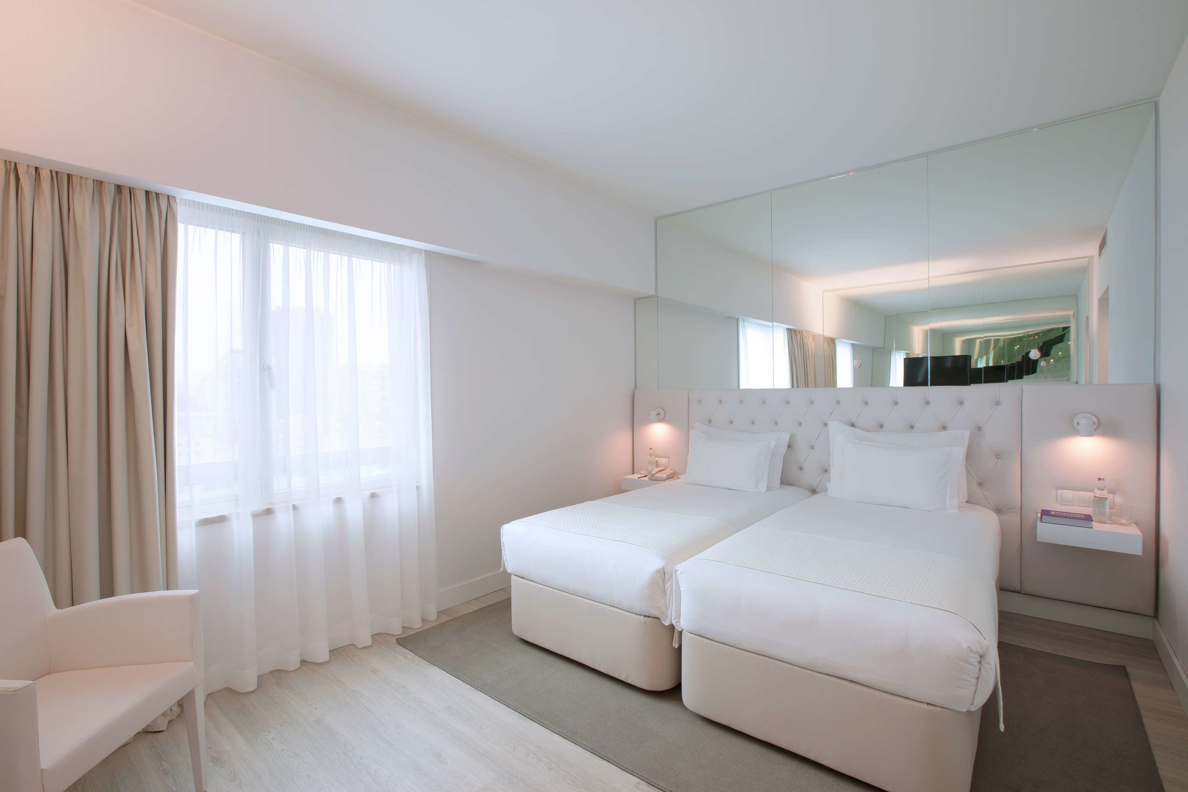A hotel room with two single beds, a white armchair, a large mirror, and a bedside table with a lamp.