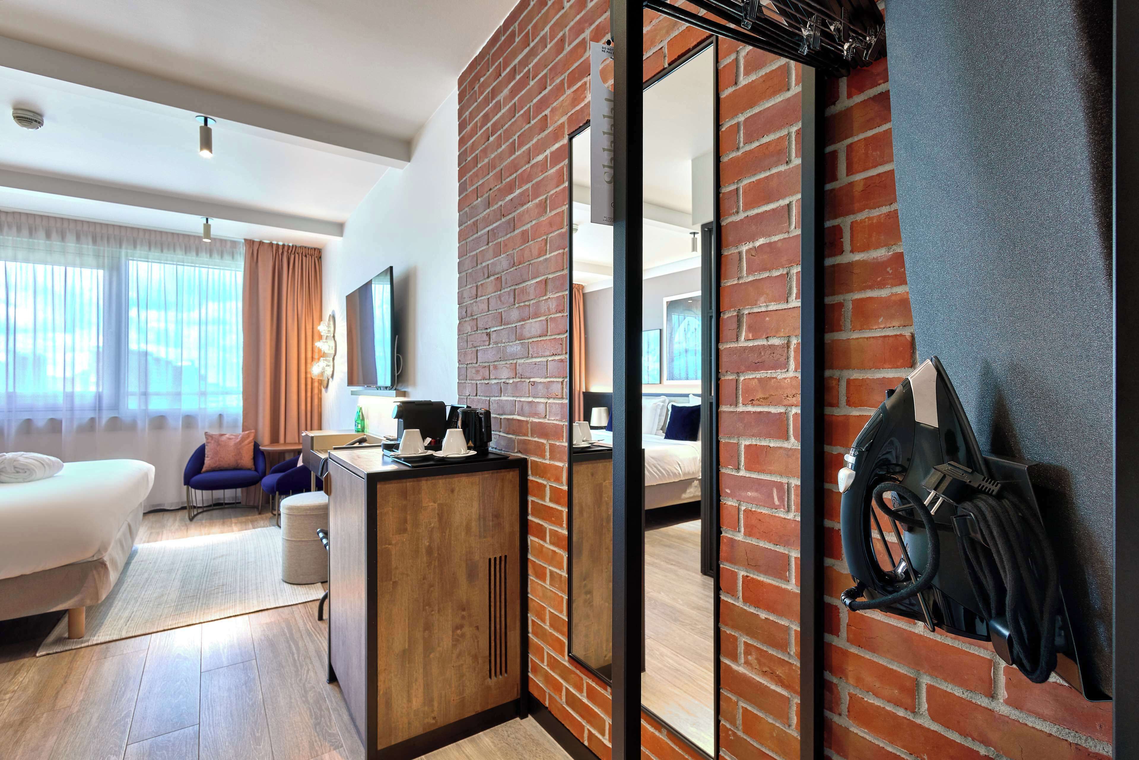 A modern hotel room with a brick accent wall, a large mirror, a bed, a desk with a coffee maker, and a charging station.