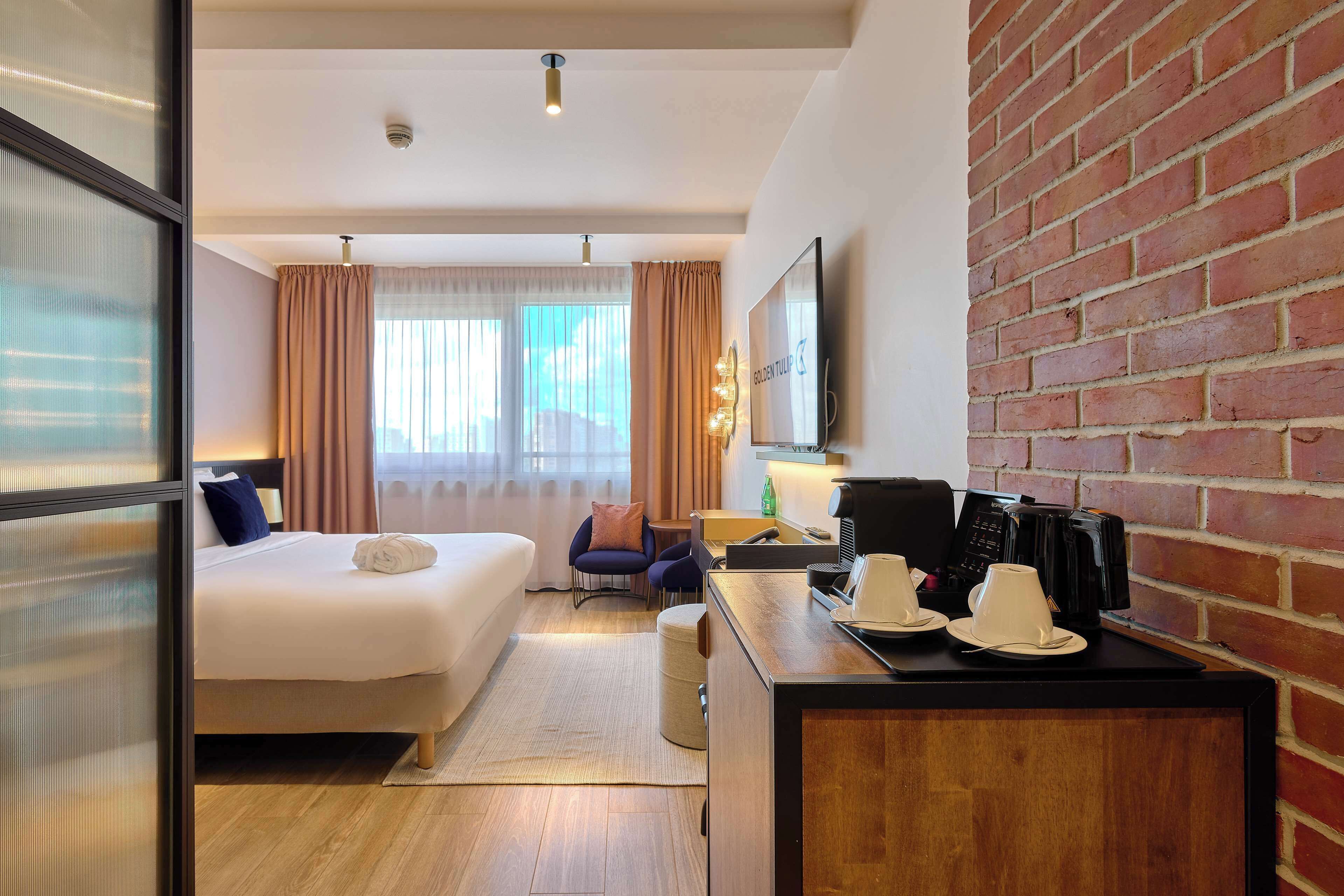 Habitación de hotel con una cama, un escritorio con tazas de café, una silla y un muro de ladrillo.
