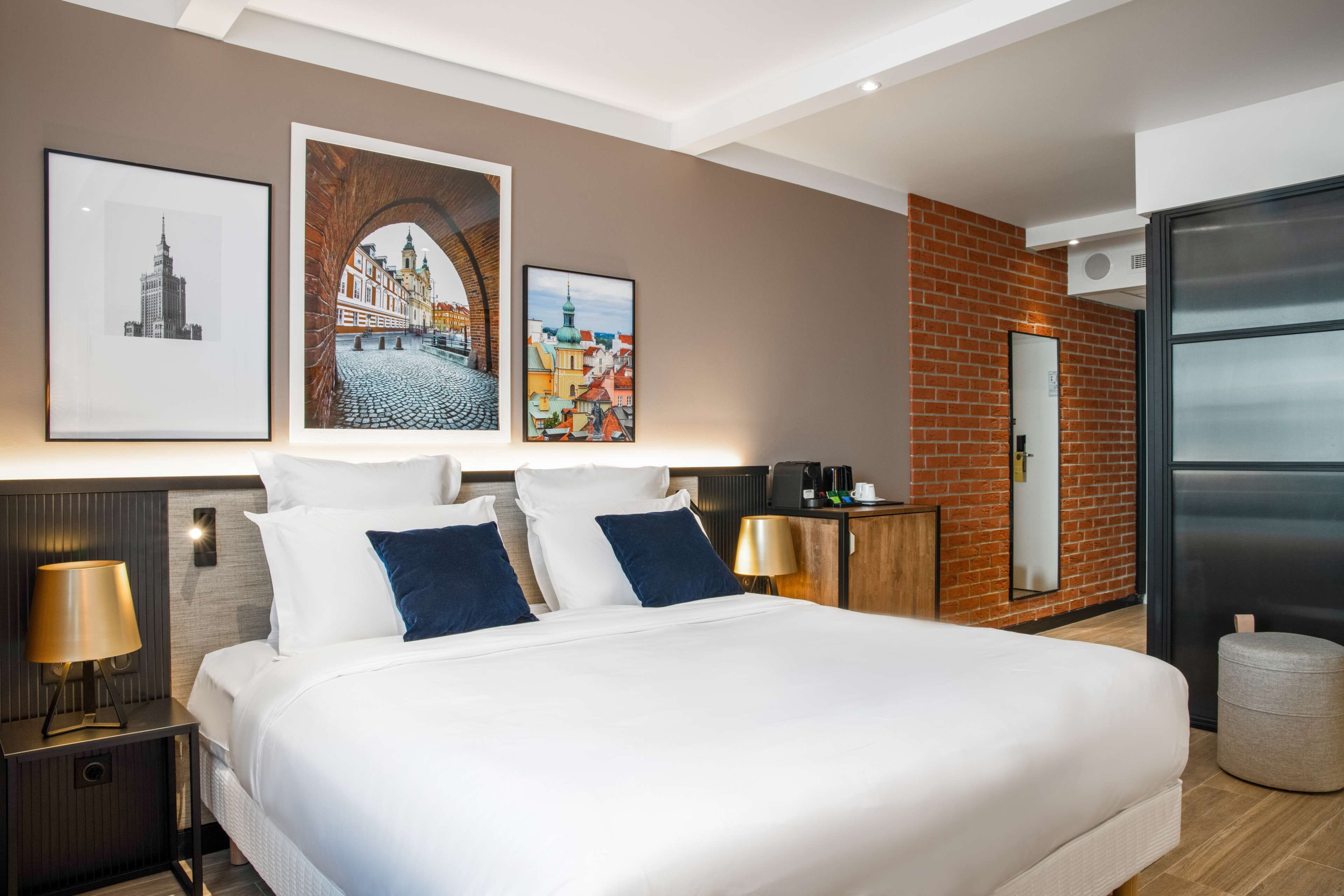 A modern hotel room with a large bed, a nightstand with a lamp, a wooden headboard, and three framed pictures on the wall.