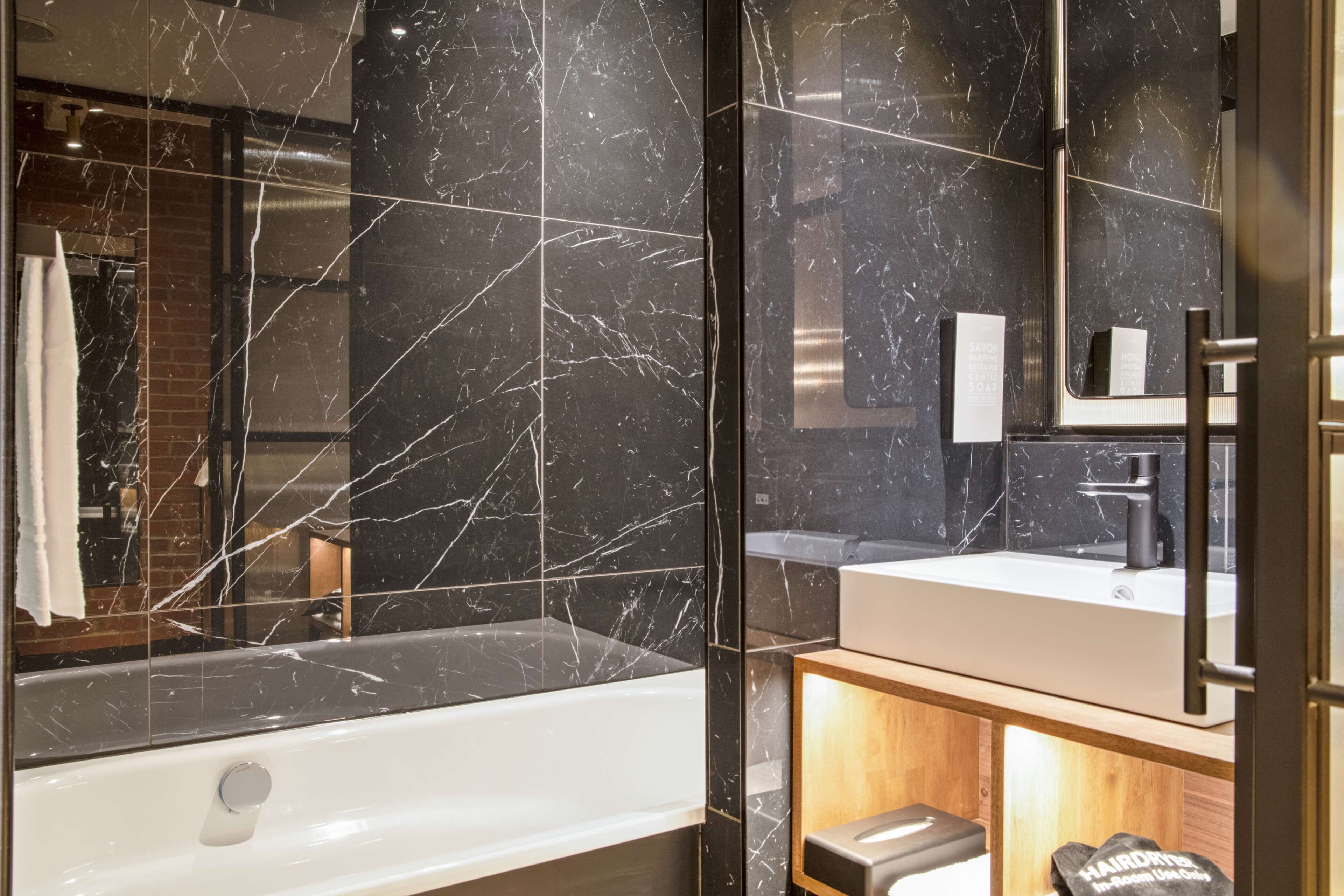 Un baño moderno con pared de azulejos de mármol, una bañera blanca y un tocador de madera con espejo.