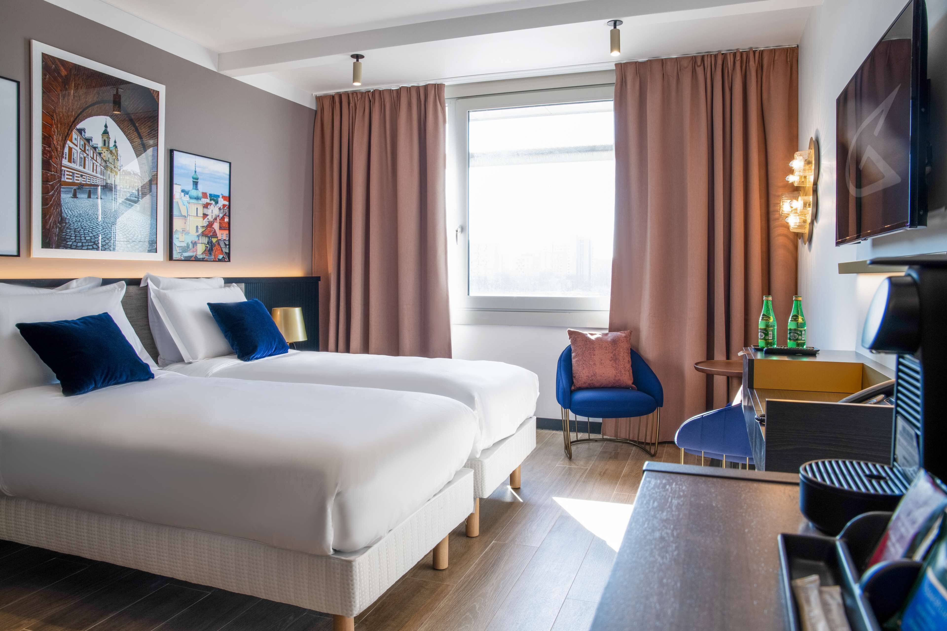 Une chambre d’hôtel avec deux lits, un bureau, une chaise, une télévision et des œuvres d’art accrochées aux murs.
