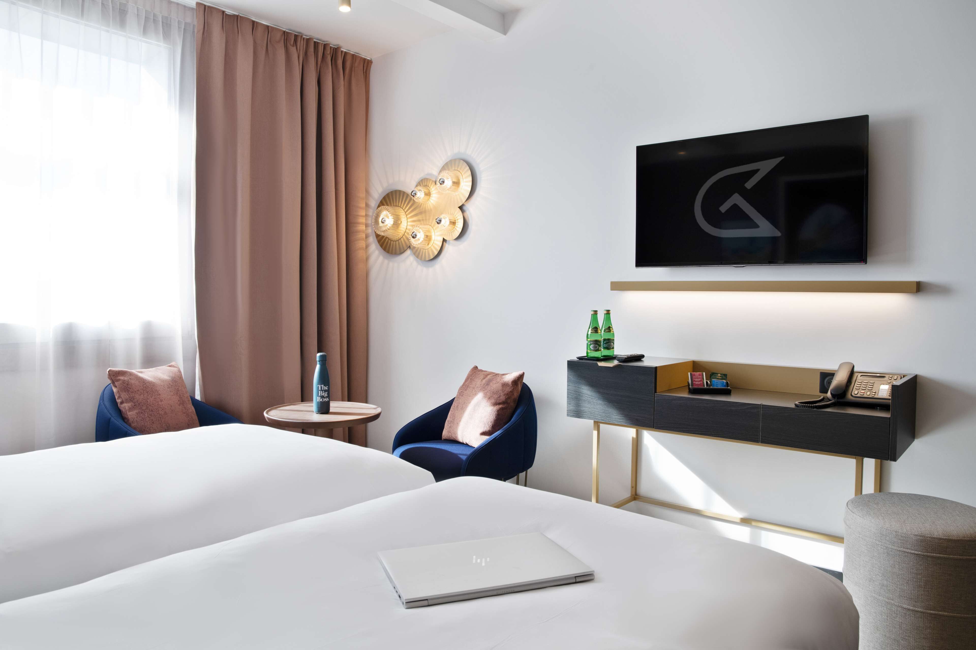A hotel room with two beds, a desk with a flat-screen TV, a small table with a lamp, and a chair.