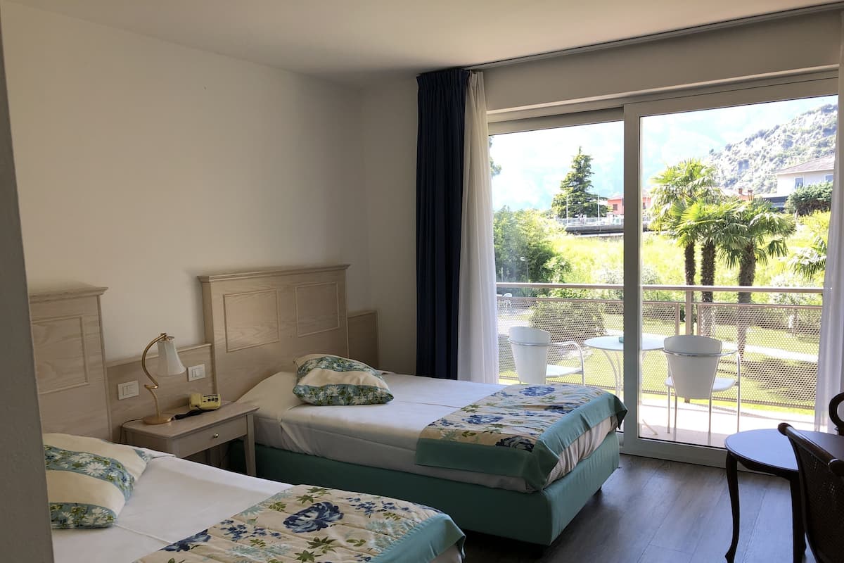 A hotel room with two beds, a balcony with outdoor seating, and a view of greenery and hills.