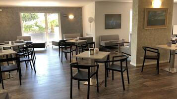 Uma sala de jantar moderna com várias mesas e cadeiras, uma janela grande com vista para o verde e um aparelho de iluminação fixado na parede.