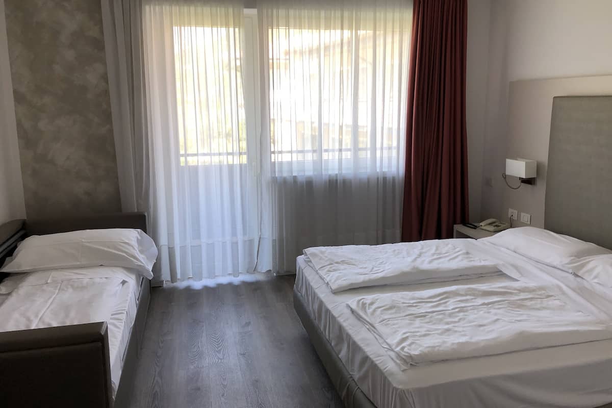 A hotel room with two single beds, a bedside table with a lamp, a window with sheer curtains, and a wall-mounted phone.