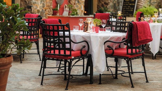 An outdoor dining area with stone walls, red upholstered chairs, and string lights. Tables are set with wine glasses and plates.