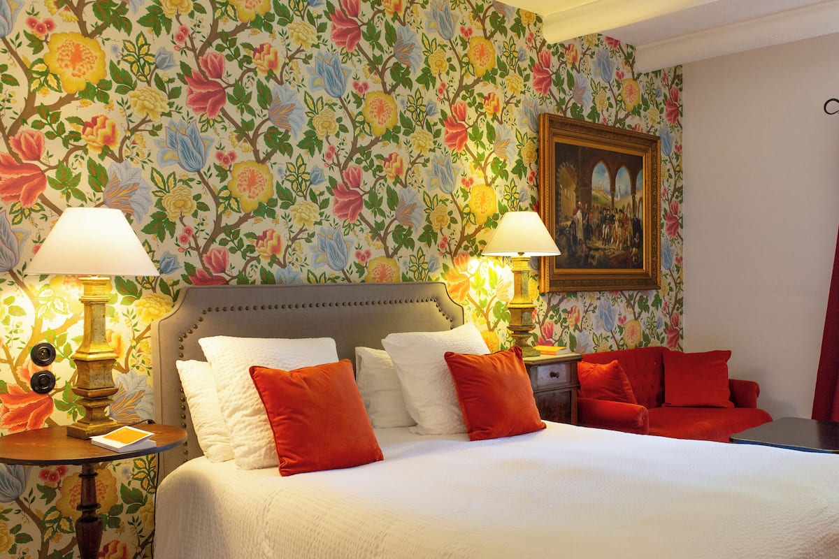 Une chambre d’hôtel avec un papier peint à motifs floraux, un lit avec des oreillers blancs et rouges, deux lampes de chevet et un tableau accroché au mur.