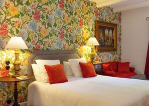 A hotel room with a floral wallpaper, a bed with white and red pillows, two bedside lamps, and a painting on the wall. - Hôtel Demeure Castel Brando (Brando)