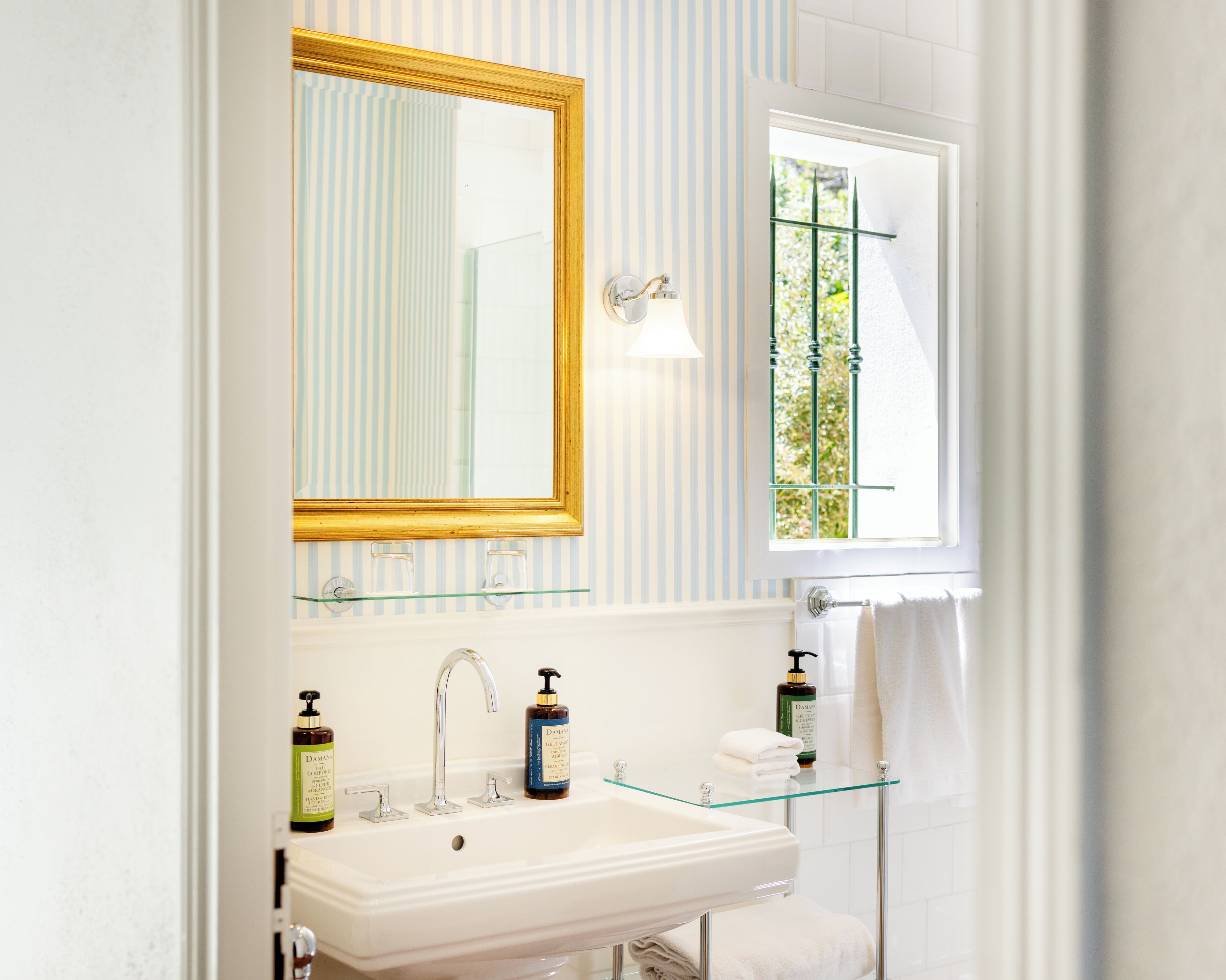 Ein Badezimmer mit einem weißen Waschbecken, zwei Shampoo-Flaschen, einem Spiegel mit goldener Rahmung und einem Fenster mit Blick auf Grünflächen.