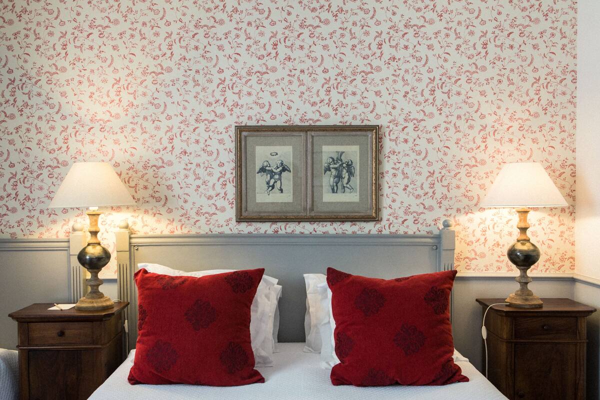 Une chambre d’hôtel avec un papier peint à motifs floraux, un lit avec des oreillers rouges, deux lampes de chevet et un tableau encadré au-dessus du lit.