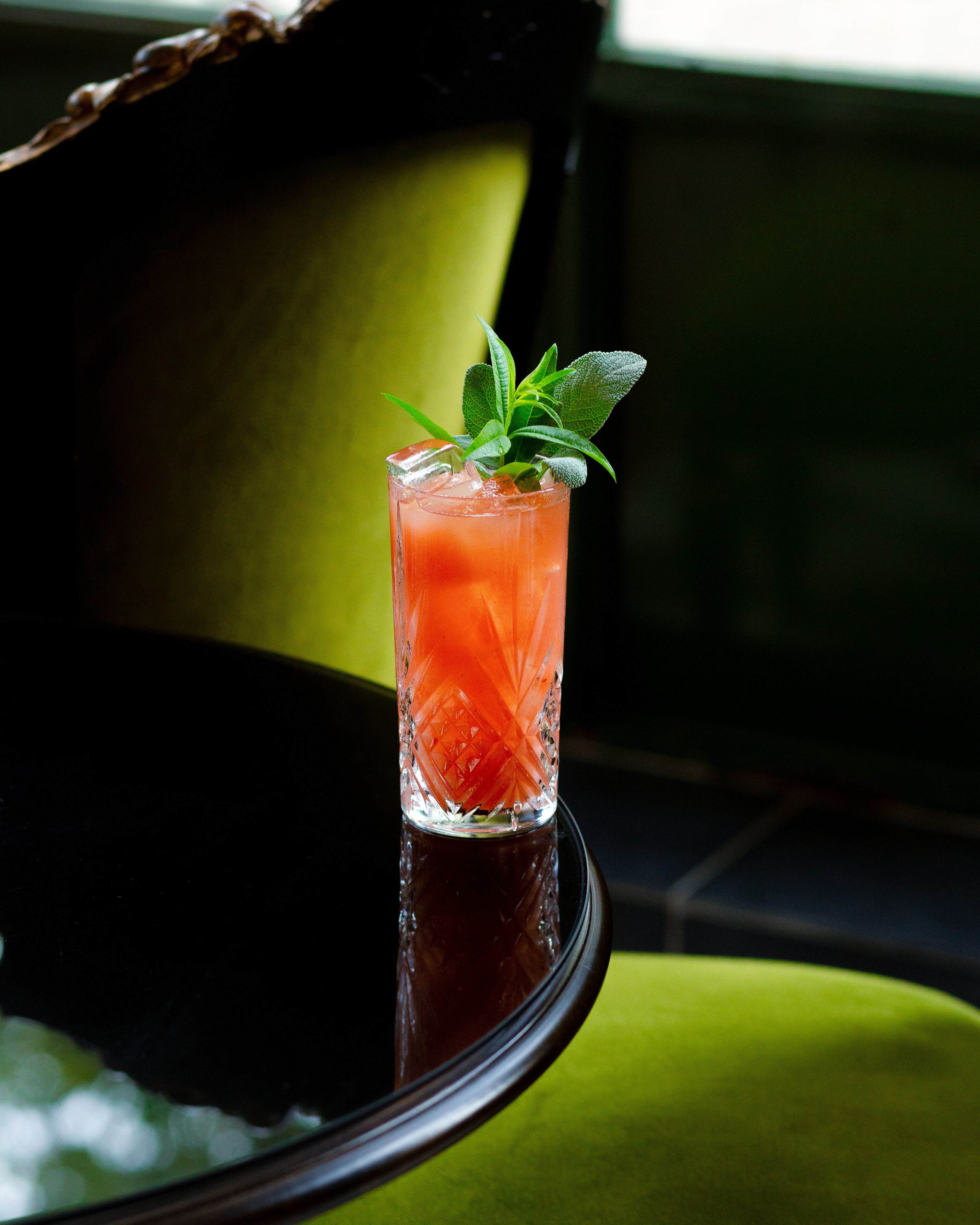 A glass of red drink with mint garnish on a reflective surface.