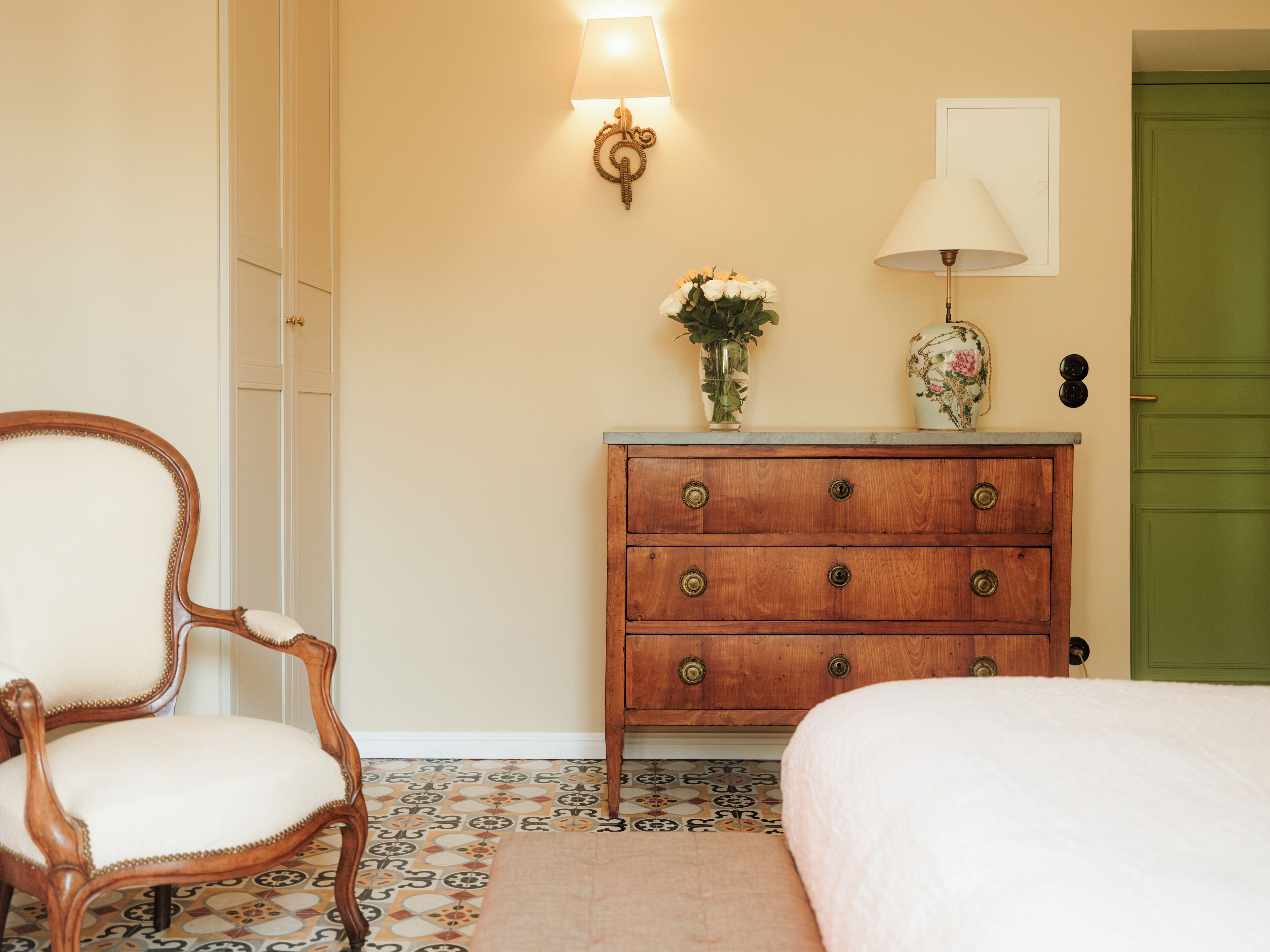 A room with a wooden dresser, a chair, a bed, a green door, and a wall-mounted lamp.