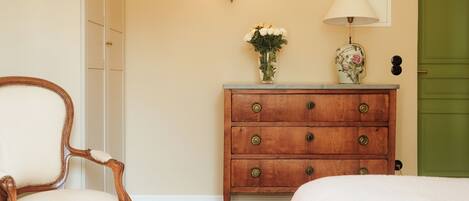 A room with a wooden dresser, a chair, a bed, a green door, and a wall-mounted lamp.