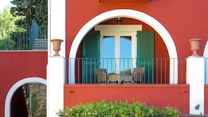 A red building with a balcony, two doors with green shutters, and a staircase leading to an upper level.