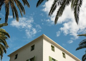 A white building with a flat roof and green shutters, surrounded by palm trees against a clear blue sky with scattered clouds. - Hôtel Demeure Castel Brando (Brando)