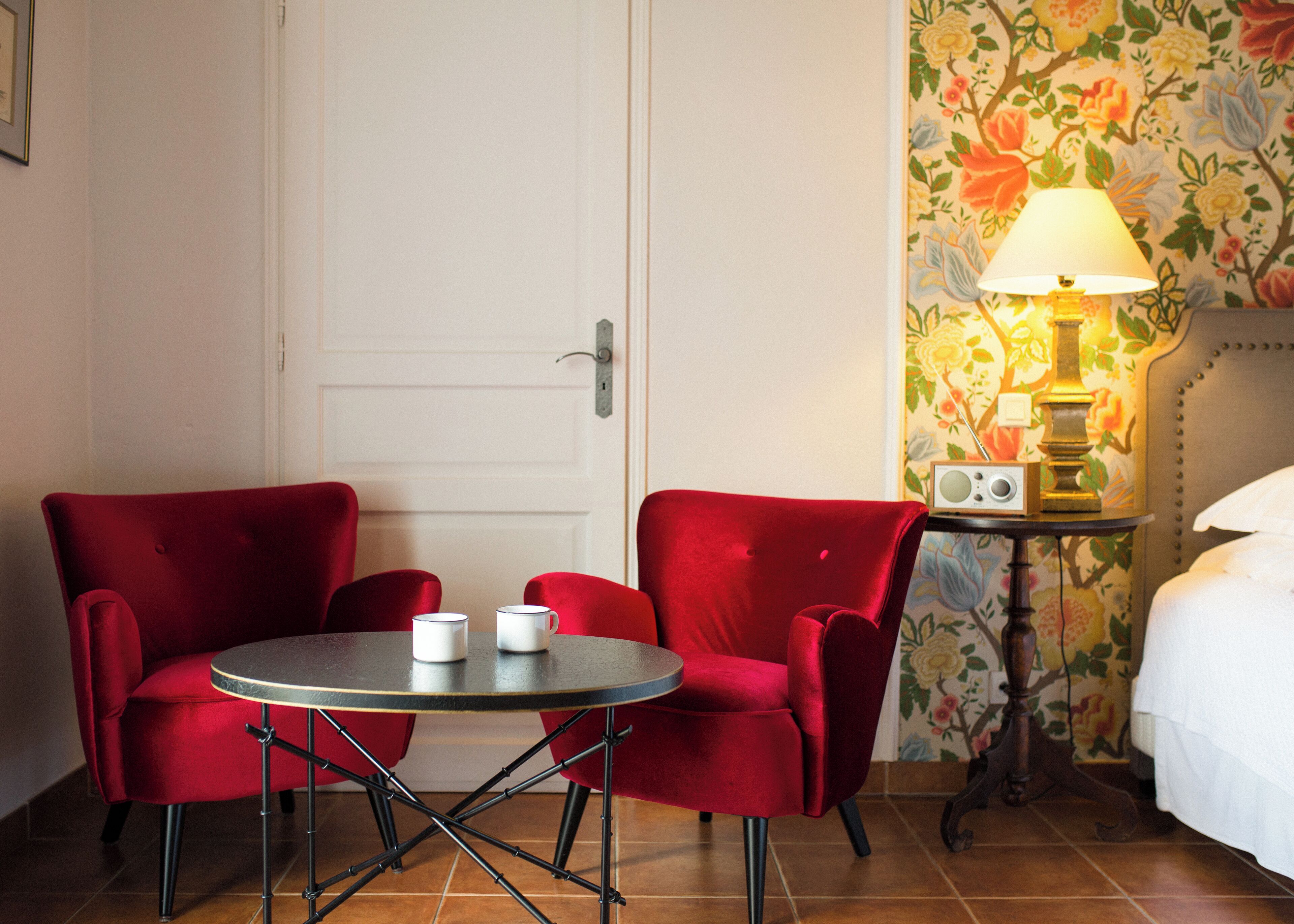 A hotel room with a red upholstered chair, a small round table with two white mugs, a lamp on a side table, and a bed with white bedding.
